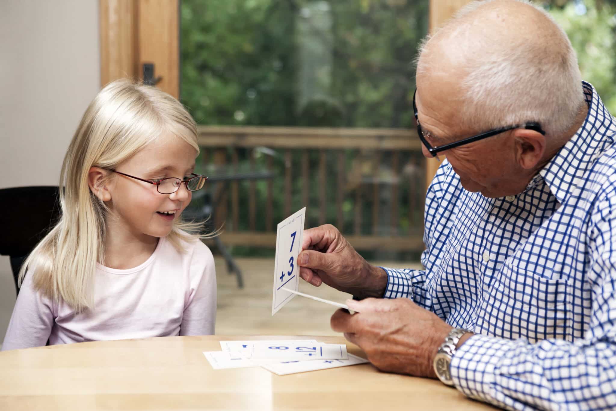 A grandfather is holding an addition flashcard for his six year old granddaughter. Seated at a dining room table with patio door in the background, the girl is smiling and engaged in the activity.